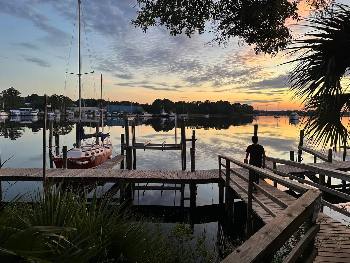 Sunset over a marina in Carrabelle, Florida, with a sailboat at the dock and a person walking the boardwalk.